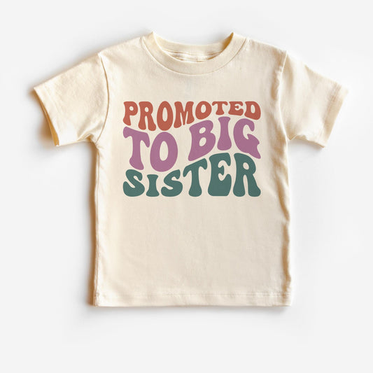 A white t-shirt with text 'Promoted to Big Sister' in colorful letters, indicating the shirt is for a young girl who has just become a big sister.