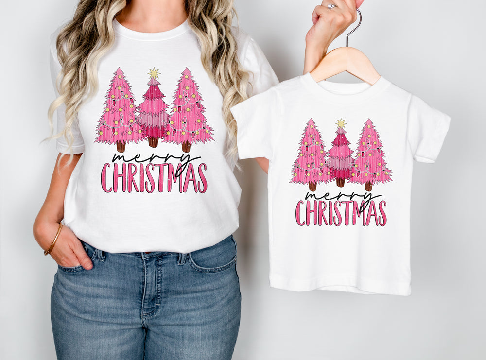 White t-shirt with pink Christmas trees and 'Merry Christmas' text, worn by a person and displayed on a hanger.