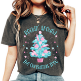 A woman wearing a Comfort Colors in pepper t-shirt with the text 'ROCKIN' AROUND THE CHRISTMAS TREE' and a colorful Christmas tree design.