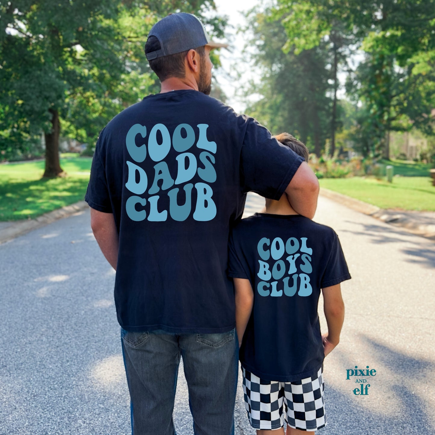 Man and child walking on a path wearing matching 'Cool Dads Club' and 'Cool Boys Club' shirts.