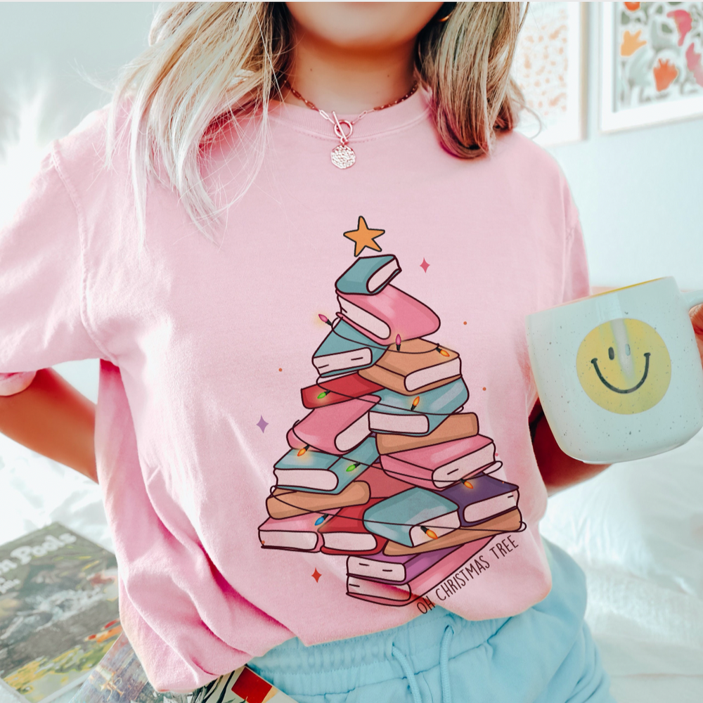 Person wearing a pink shirt with a book-themed Christmas tree design, holding a mug.