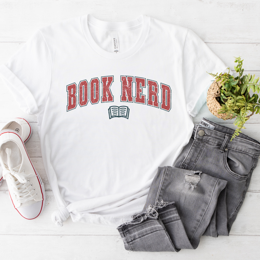 White t-shirt with 'BOOK NERD' text in red and blue on the front, displayed flat with a pair of gray jeans and white sneakers to the side.