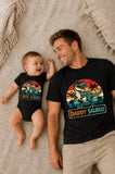 Man and baby wearing matching 'Daddy Saurus' and 'Mini Dinosaur' shirts on a textured surface.