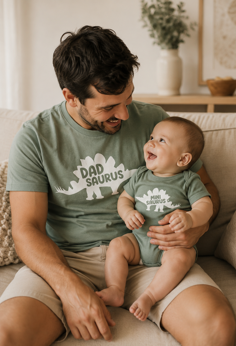 Man and baby wearing matching 'Dad Saurus' shirts sitting on a couch.