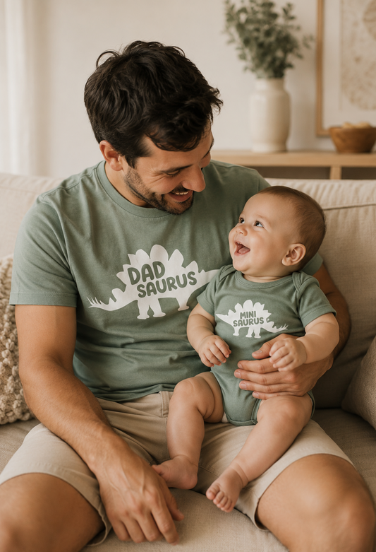 Man and baby wearing matching 'Dad Saurus' shirts sitting on a couch.