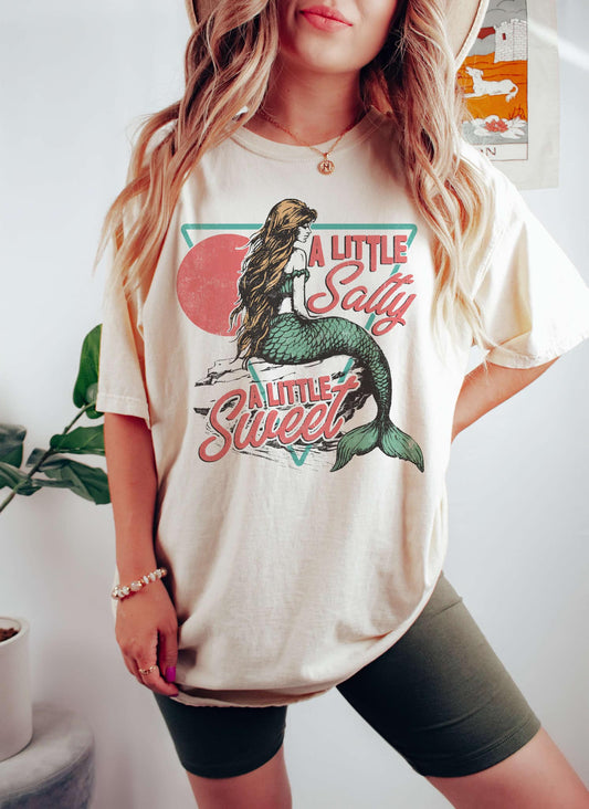 A woman wearing an ivory colored T-shirt with a graphic print of a mermaid sitting on a surface, with the text 'A Little Salty' above it.