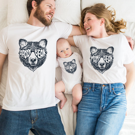 A family of three, including a baby, a toddler, and an adult, all wearing white t-shirts with a black bear design and the text 'Daddy Bear'. They are lying on a bed, smiling and posing for the camera.