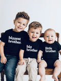 three brothers sitting on a bench wearing black shirts with the print on each biggest brother, big brother and little brother.