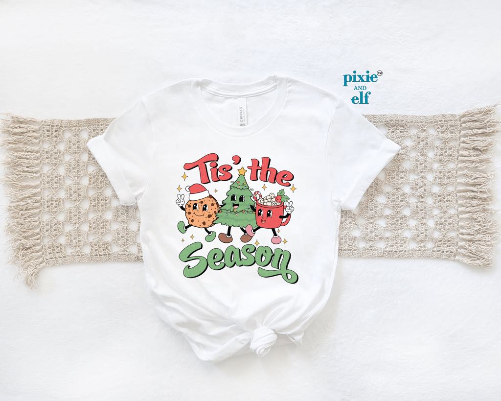 A white t-shirt with a Christmas themed graphic that says 'tis the season' and features cartoon images of Christmas cookies, christmas tree and cup of hot coco.