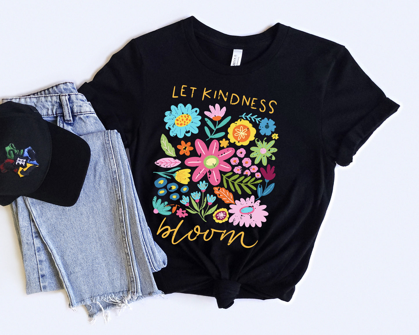 Black t-shirt with colorful floral design and 'Let Kindness Bloom' text, paired with a black cap and jeans on a white background.
