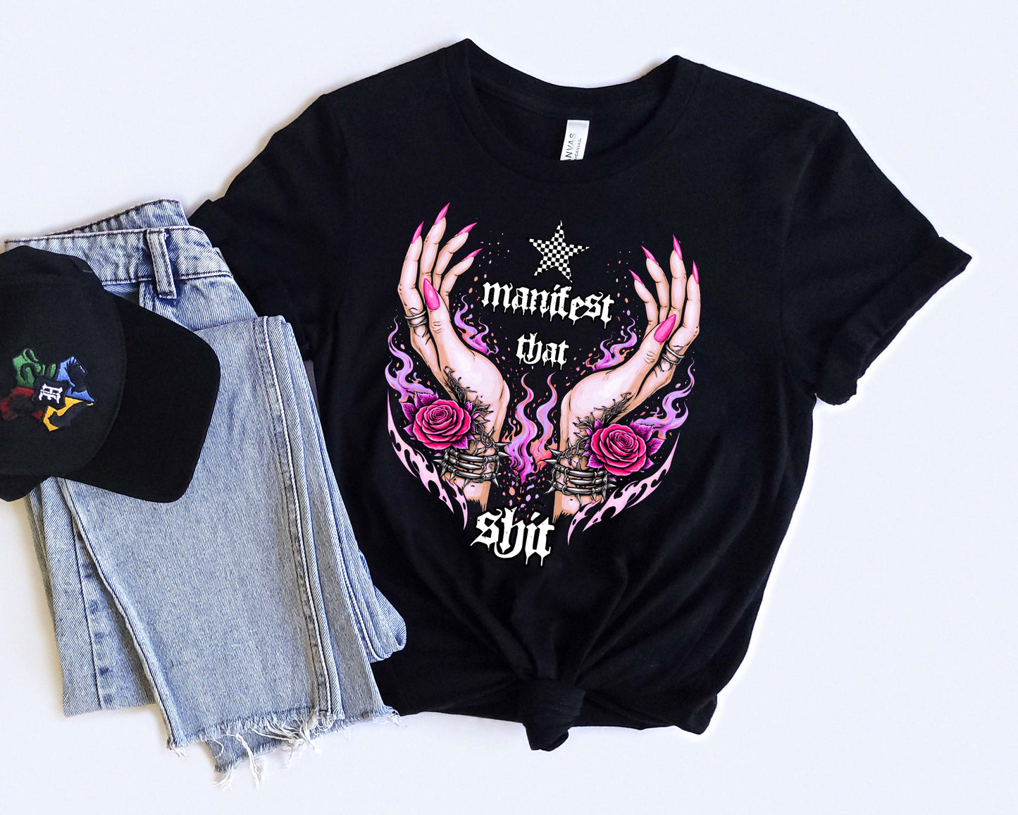 Black t-shirt with graphic design and text "Manifest that Shit", paired with denim shorts and a black cap on a light background