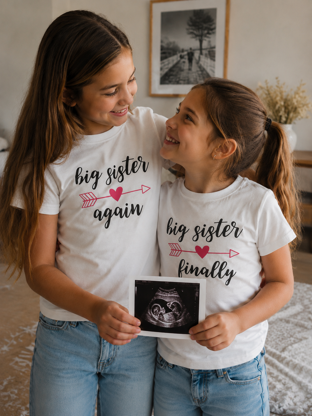Two sisters wearing 'big sister again' and 'big sister finally' shirts, holding an ultrasound photo.