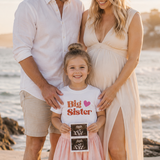 Family of three on a beach with a pregnant woman and a young girl holding an ultrasound photo.