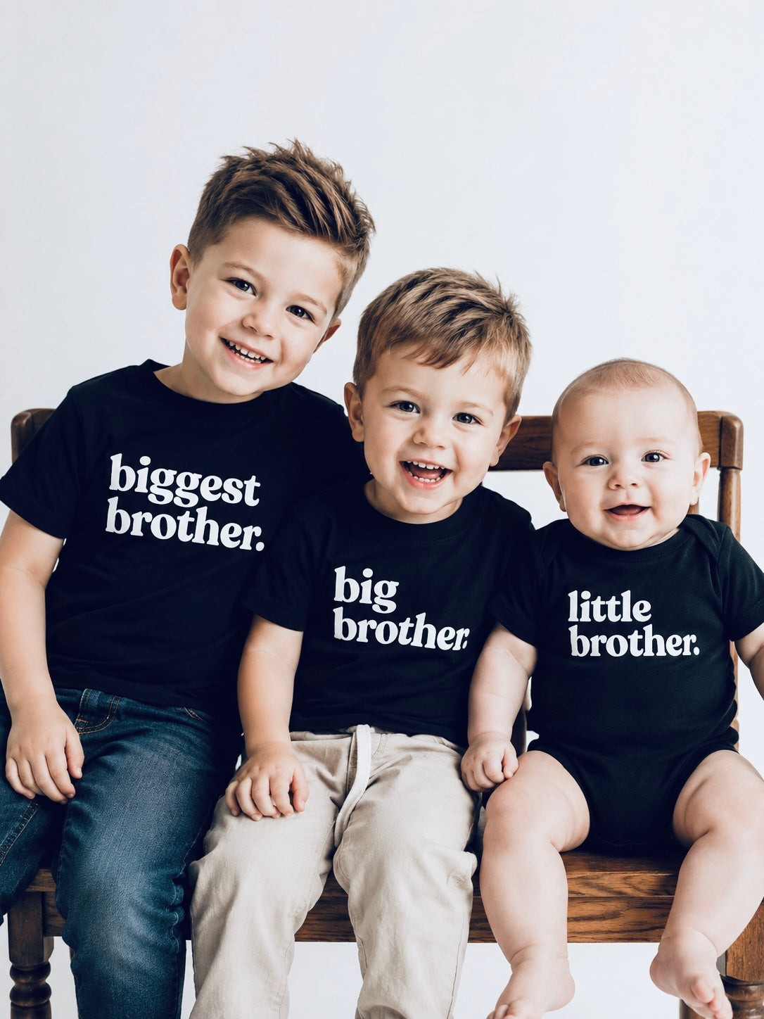 Three children wearing black shirts labeled 'biggest brother', 'big brother', and 'little brother' sitting on a chair.