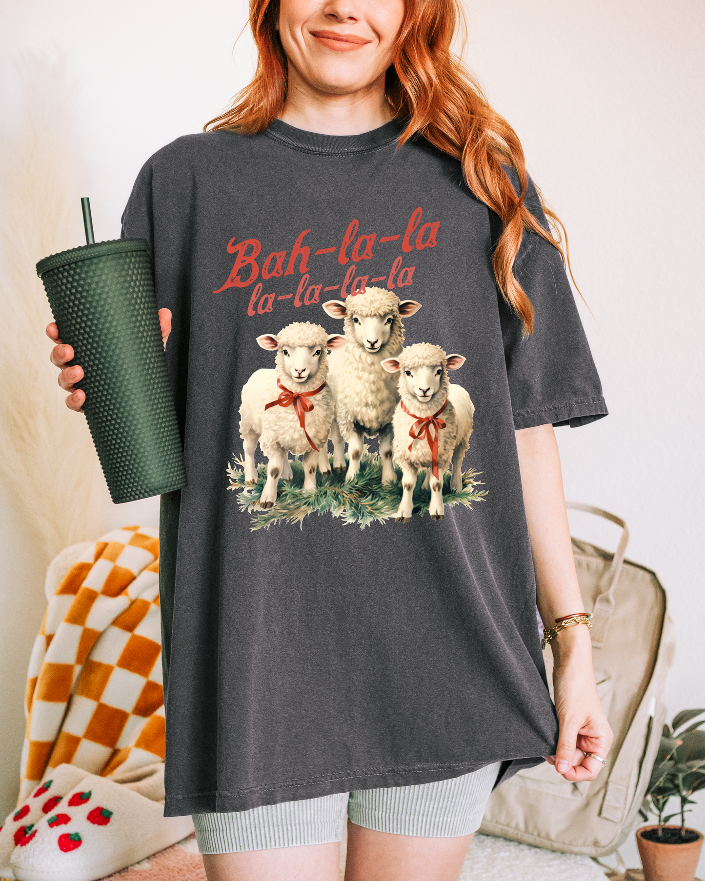 Person wearing a dark gray t-shirt with sheep graphic and 'Bah-la-la' text, holding a green tumbler.