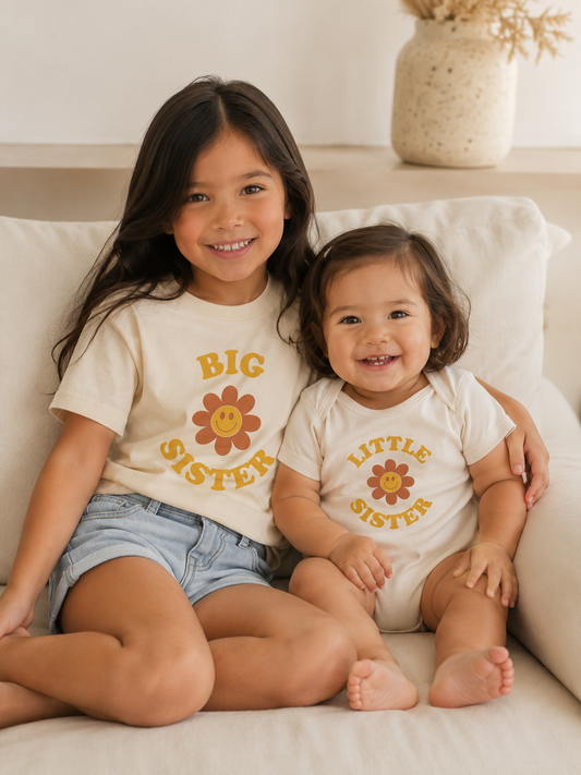 big sister and little sister sits in the couch and wears a big sister shirt little sister bodysuit  with a flower accent.