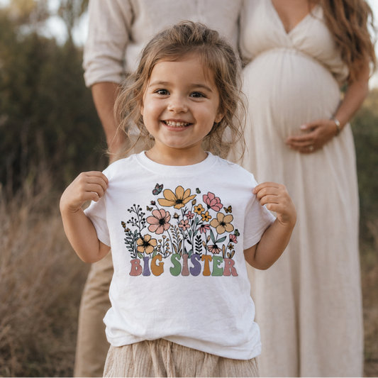 Family of three outdoors with a young girl wearing a 'Big Sister' shirt.